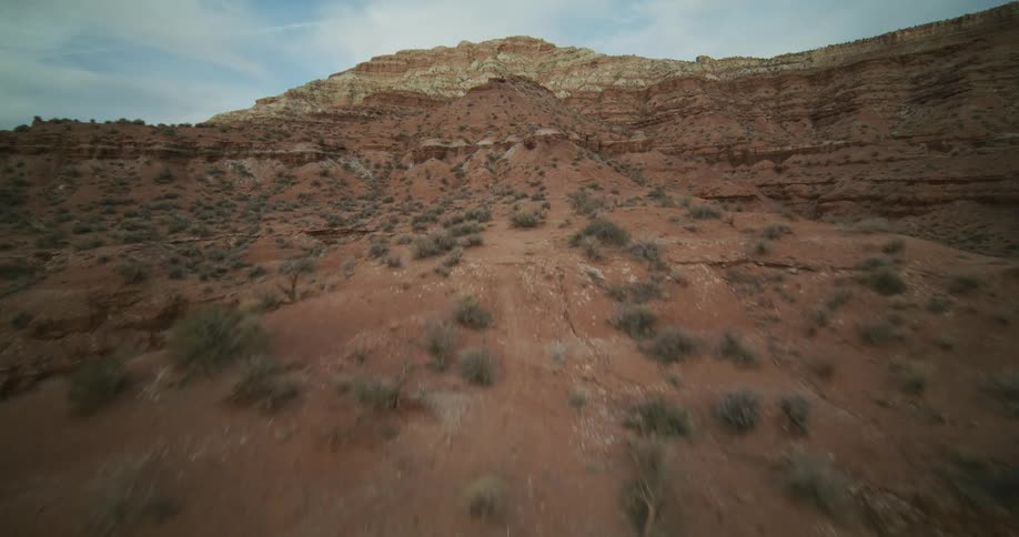 An immersive aerial view glides up a textured red earth hillside towards a vast, layered desert butte