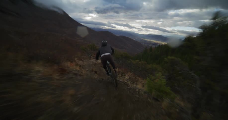 Dynamic follow-shot of a mountain biker speeding down a rugged trail in a moody, overcast mountain landscape