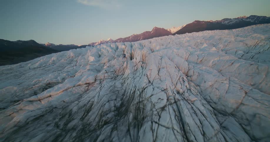 Epic aerial view reveals a helicopter landing on a vast, icy glacier amidst a stunning Alaskan mountain range at sunset