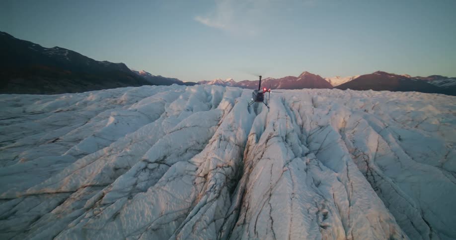 A sightseeing helicopter lands on the fissured surface of a vast glacier at dusk, surrounded by a dramatic mountain range