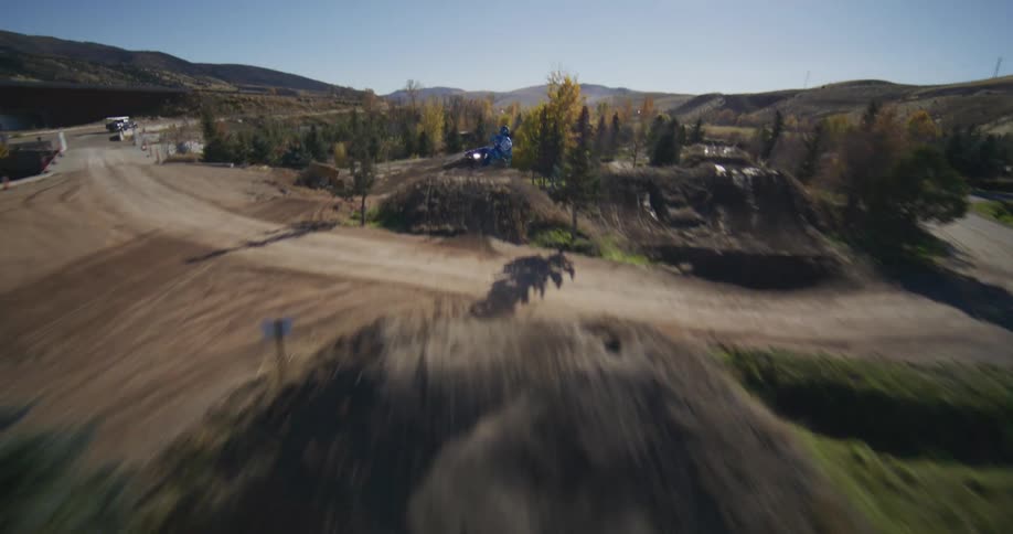 Dynamic aerial view follows a motocross rider in blue racing gear as they speed along a dirt track and launch off large jumps on a sunny day