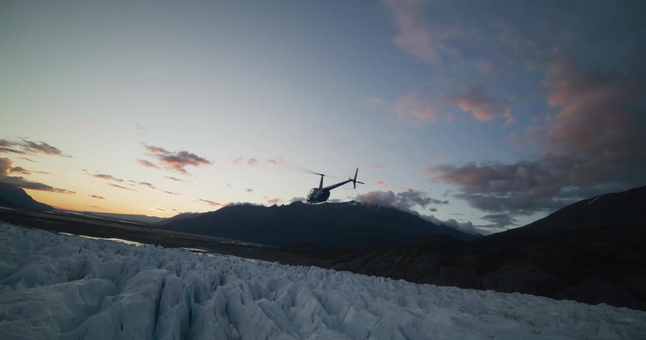 A helicopter slowly flies away over a massive glacier during a scenic sunset in the mountains
