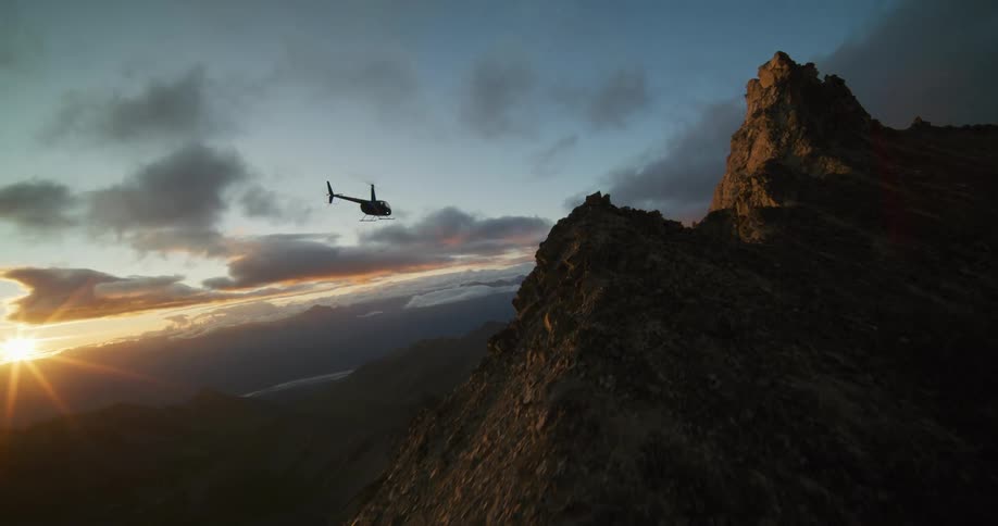 A helicopter takes off from a dramatic mountain crest at sunrise and flies over the vast, epic landscape