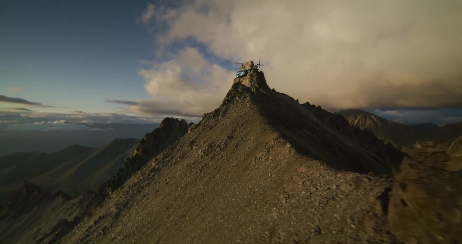 A stunning aerial shot follows a helicopter flying along a jagged mountain ridge before disappearing behind the peak during a golden sunset
