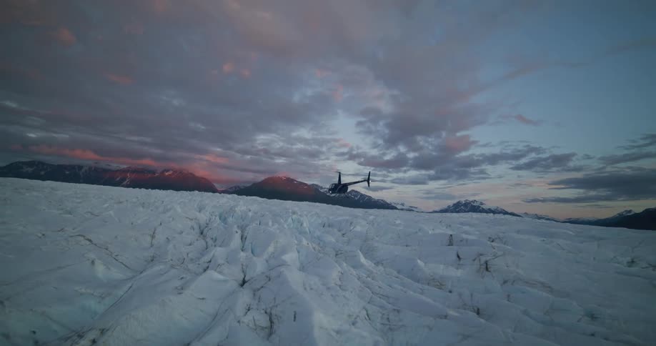 A helicopter takes off from a massive, crevasse-filled glacier and flies into the colorful sunset sky over the mountains
