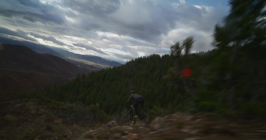 A fast-paced aerial follows a mountain biker riding a scenic and narrow ridgeline trail under a dramatic, cloudy sky