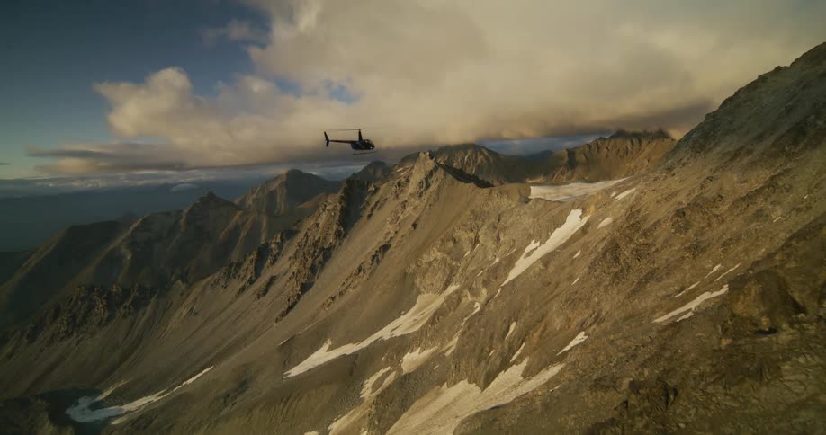 Cinematic slow-motion aerial footage captures a helicopter flying over a rugged mountain range at sunset