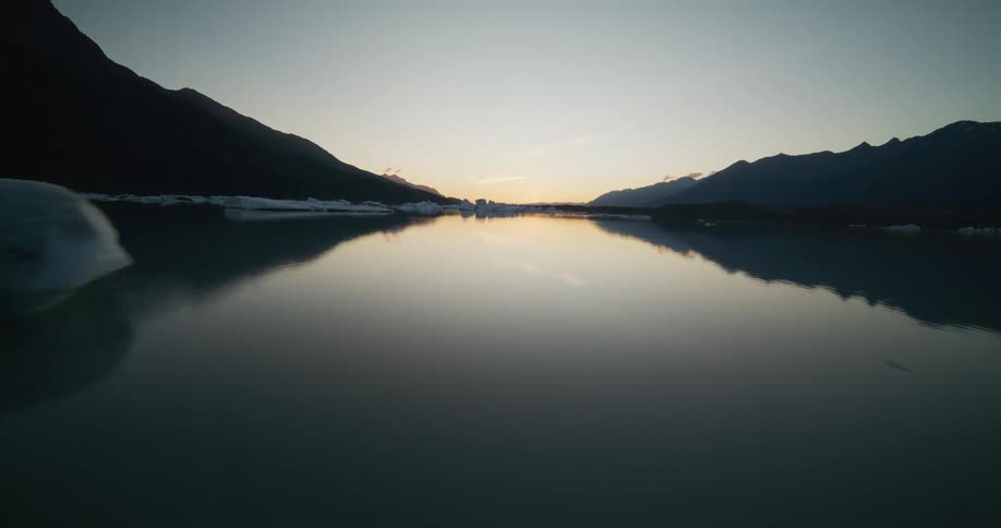 A slow aerial flyover of melting icebergs in a tranquil glacial lake at sunset, surrounded by majestic mountains