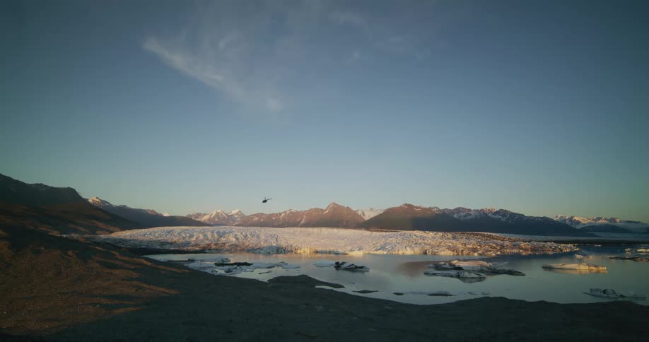 A helicopter flies over a vast glacier and iceberg-filled lake with a mountain range in the background at sunset