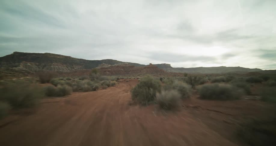 A dizzying point-of-view shot speeds along a winding dirt road through a vast red rock desert canyon under an overcast sky