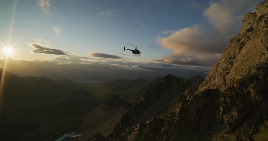 An epic aerial shot follows a helicopter as it flies along a dramatic mountain ridge and soars into the golden sunrise over a vast valley