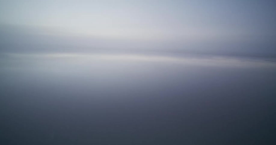 An ethereal aerial journey over a foggy, reflective sea, rising through the clouds to reveal the serene sky at dawn
