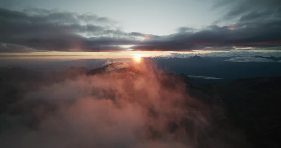 An epic aerial shot flies back through pink clouds revealing a stunning sunrise over a vast mountain range