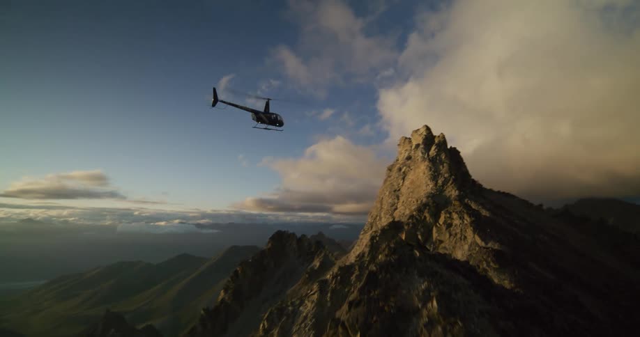 A helicopter flies away over a dramatic mountain ridge into the vast, golden-lit landscape at sunrise