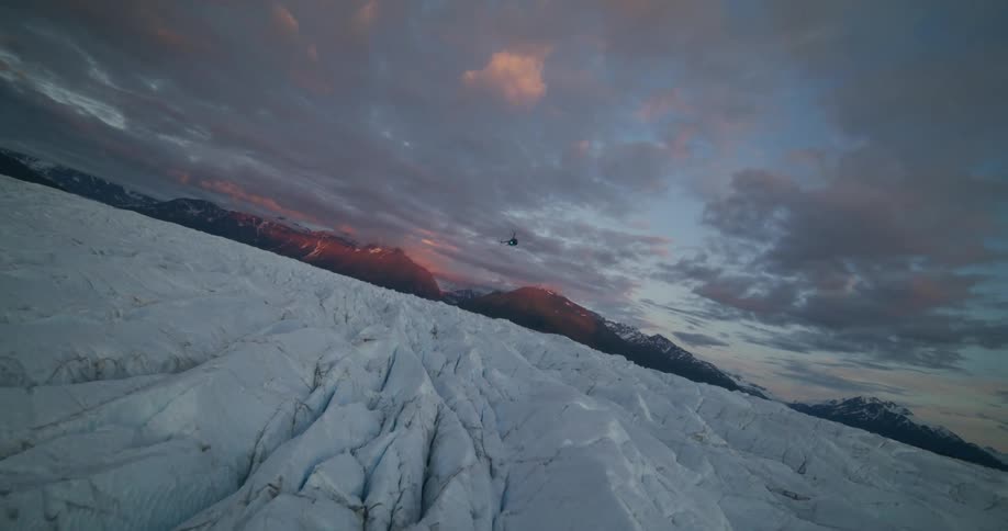 A dramatic aerial shot follows a helicopter flying over a vast, icy glacier as the sun sets behind distant mountains