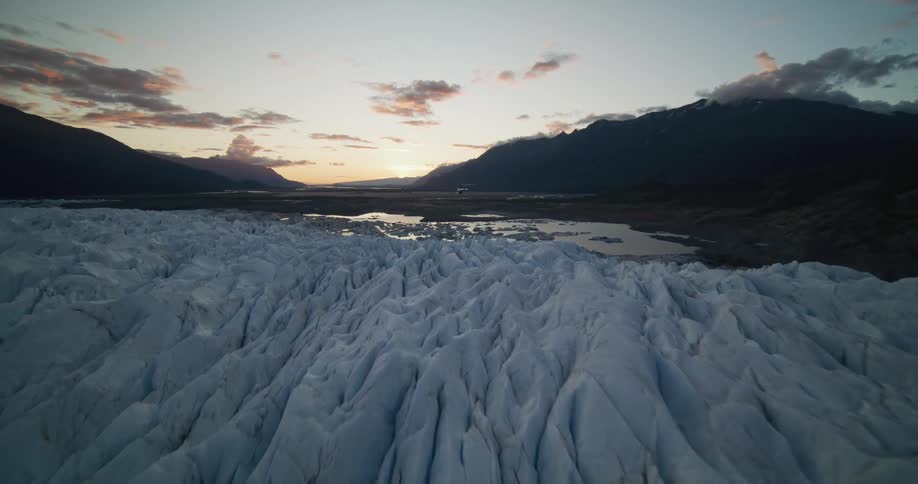 A stunning aerial flight low over the jagged, crevassed surface of a vast glacier towards a mountain valley and glacial lake at sunset