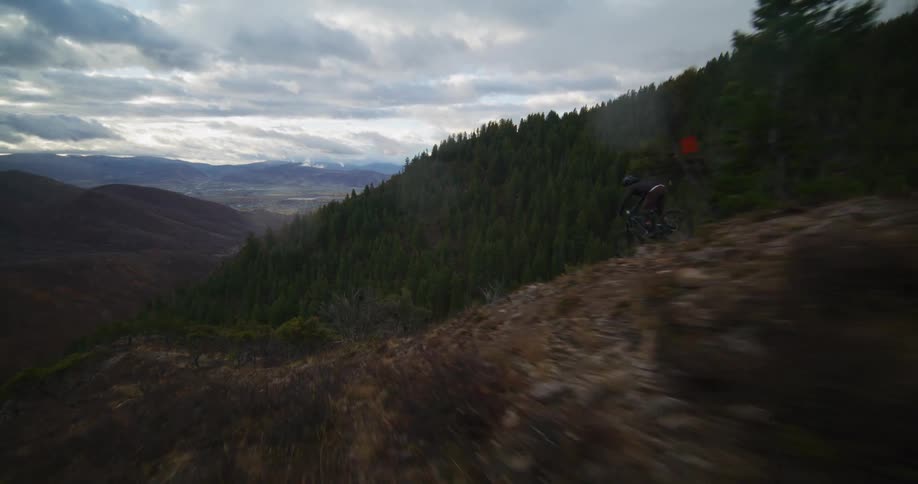 An exhilarating aerial follow shot tracks a mountain biker skillfully descending a rugged trail in a vast, moody landscape
