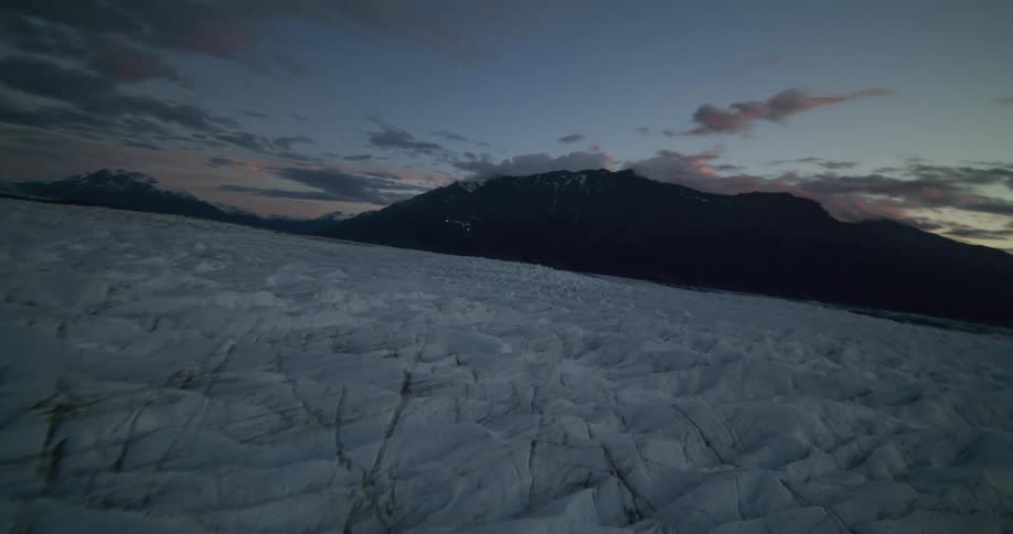 A helicopter flies over a vast, icy glacier surrounded by mountains during a dramatic dusk