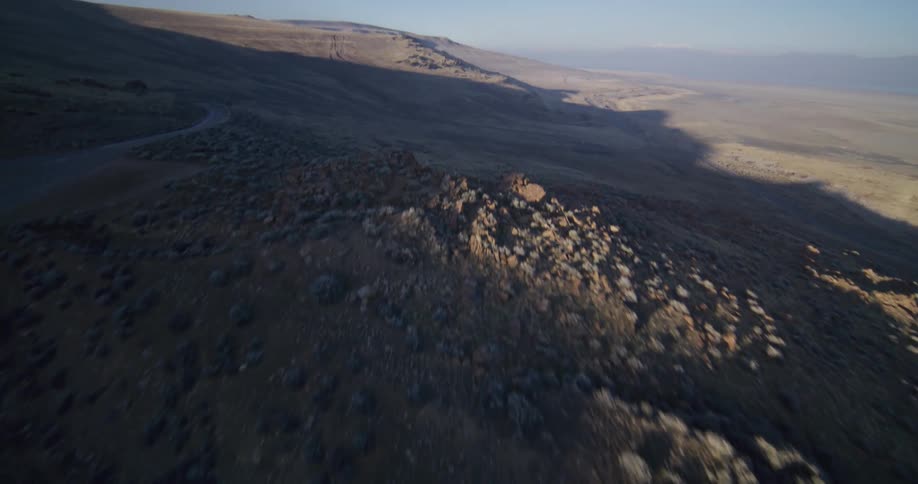 A dramatic aerial shot flies over a rocky hillside at sunrise, revealing a vast, arid landscape and a distant winding road