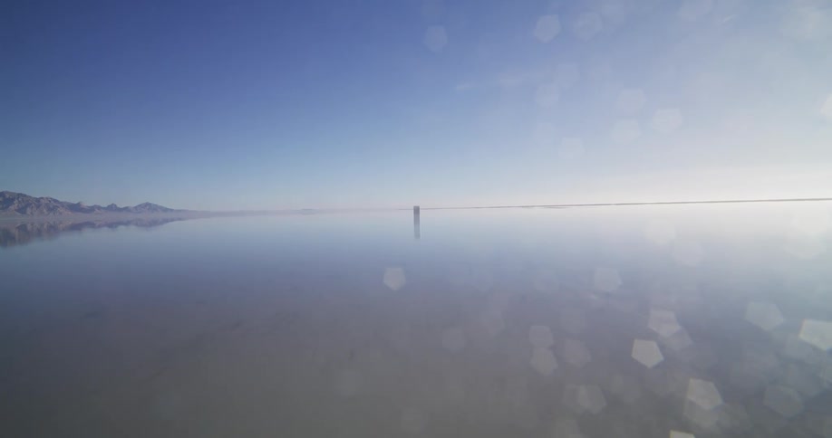 A surreal aerial journey towards a mysterious metal monolith standing alone on a vast, reflective salt flat under a clear blue sky