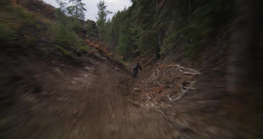 An aerial view tracks a mountain biker as they expertly navigate a steep, winding trail through a dense, rugged forest