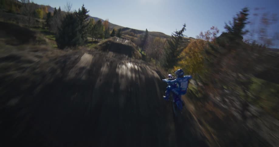 An FPV drone chases a motocross rider in blue gear as they race around a dirt track in the countryside on a sunny day