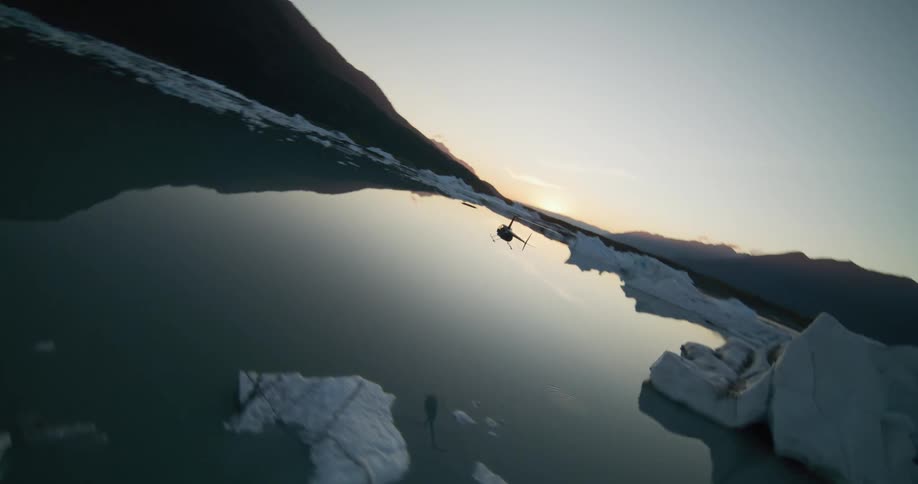 A helicopter soars over a reflective glacial lake filled with icebergs as the sun sets behind distant mountains