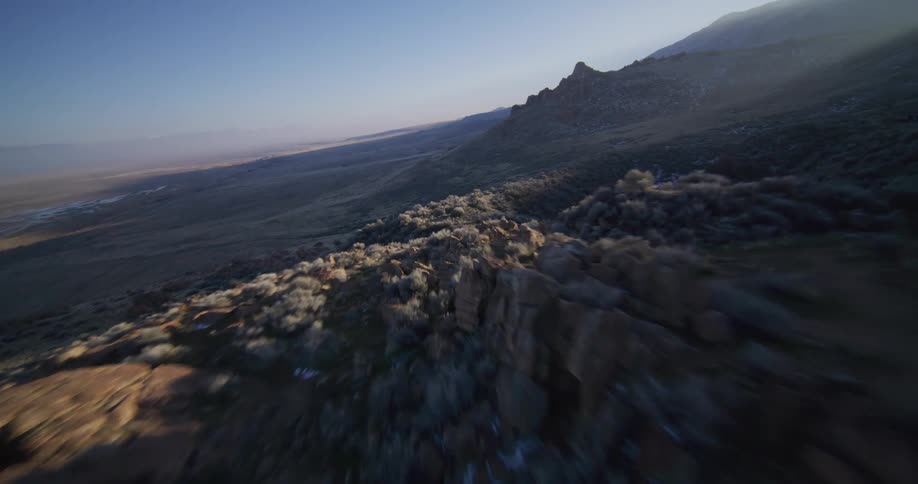Exhilarating FPV drone flight dives down a rugged, sunlit mountainside ridge covered in desert brush