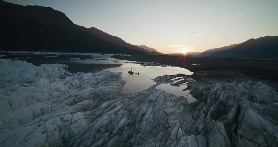 A helicopter hovers over a serene glacial lake at sunset, surrounded by vast, rugged glaciers and distant mountains
