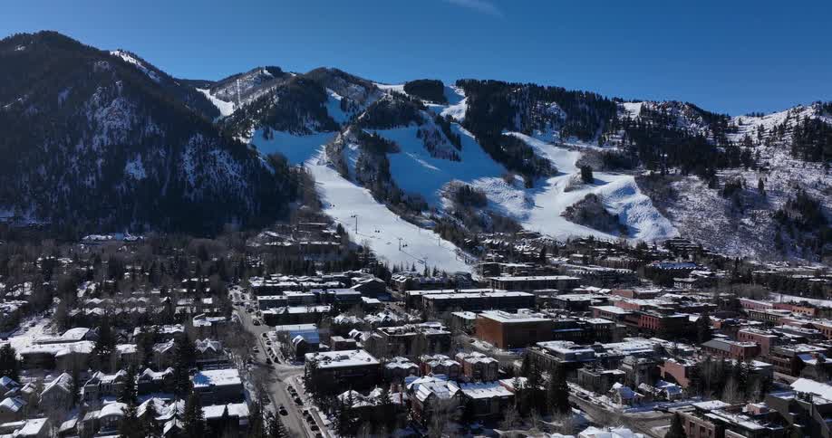 A slow aerial pullback reveals a picturesque ski resort town nestled at the base of a snow-covered mountain on a sunny winter day