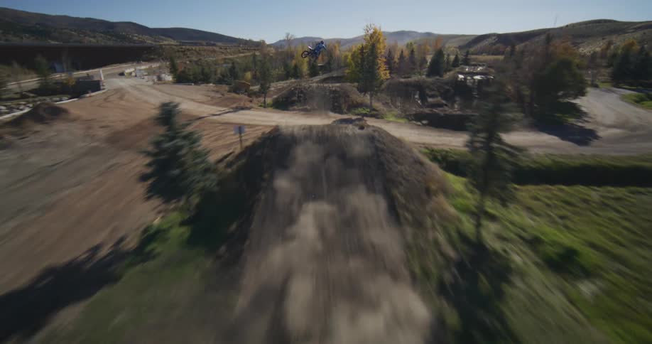 An aerial view descends to follow a motocross rider as they speed along a winding dirt track and launch over a large jump