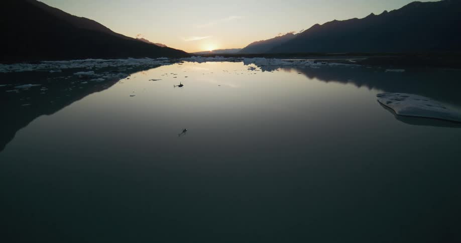 An FPV drone pursues a helicopter flying low over a still, icy glacial lake surrounded by mountains at sunset