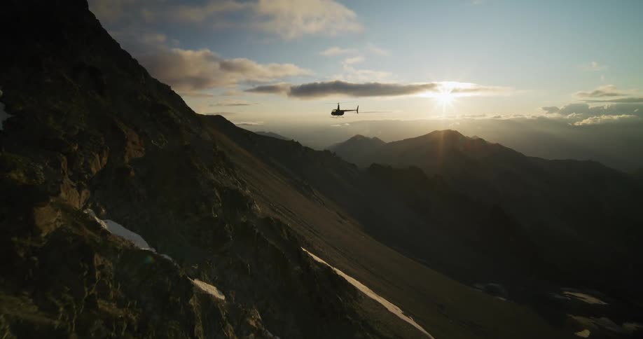 An epic aerial view follows a helicopter as it flies away from a dramatic, rocky mountainside into the sunset