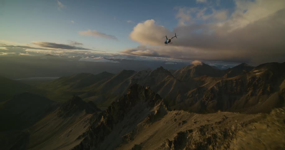 A helicopter takes off from a rugged mountain peak and flies into the stunning sunset over a vast mountain range