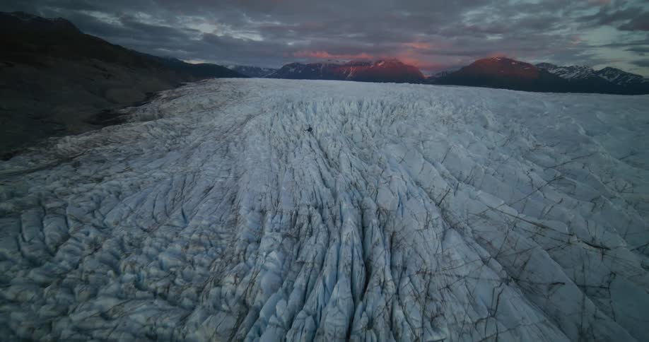 A dramatic aerial view shows a helicopter flying low over the vast, crevasse-filled surface of a remote Alaskan glacier at sunset