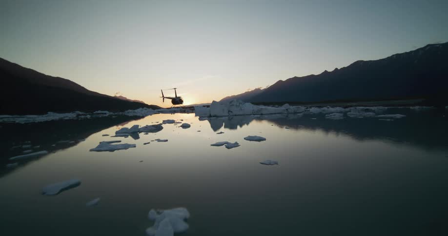 A stunning aerial view captures a helicopter flying low and landing beside a reflective glacial lake filled with icebergs during a serene sunset