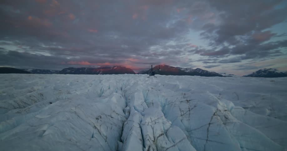 A helicopter flies away over a vast, cracked glacier during a beautiful sunset in the mountains
