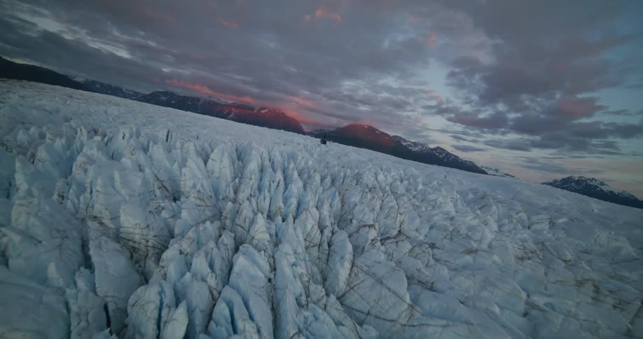 A dramatic aerial shot follows a helicopter flying low over a vast, crevasse-filled glacier at sunset with mountains in the background