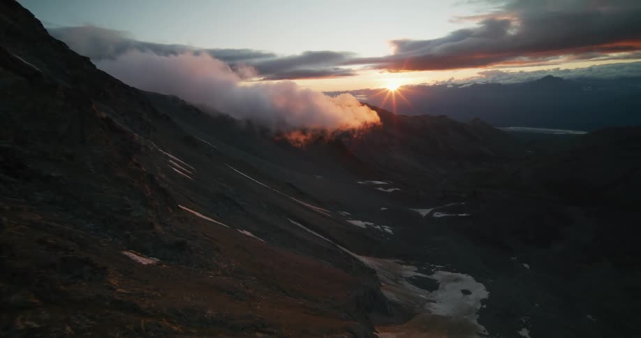 Dramatic aerial footage reveals a golden sunset over a vast mountain range, with glowing clouds drifting through the valley