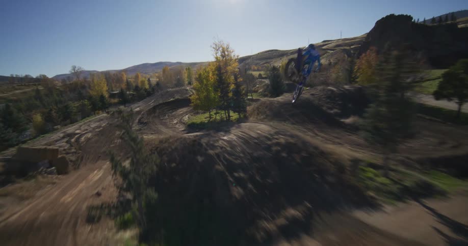 An immersive FPV drone follows a motocross rider performing tricks and jumps on a sunny rural dirt track