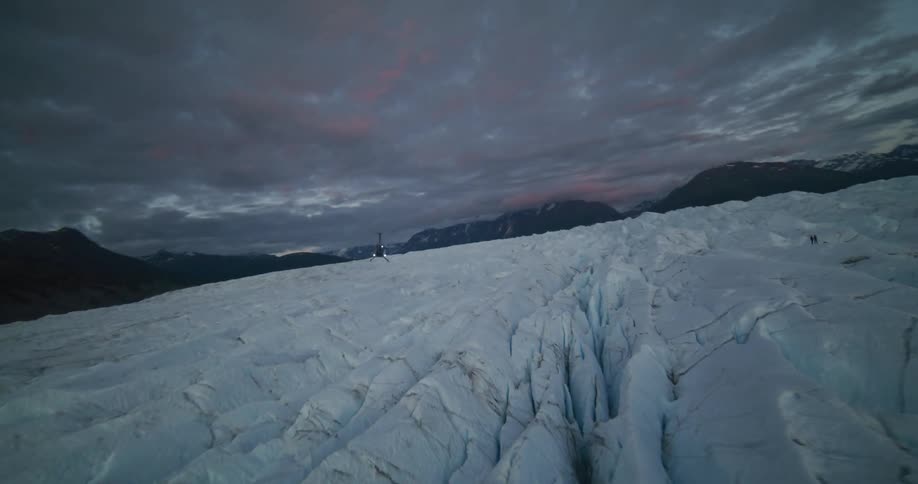 A helicopter with flashing lights lands on a vast, cracked glacier under a dramatic sky at twilight