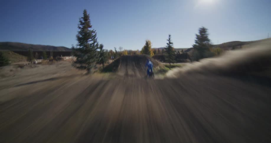 Exciting FPV drone shot follows a motocross rider in slow motion as they launch off a large dirt jump on a sunny day