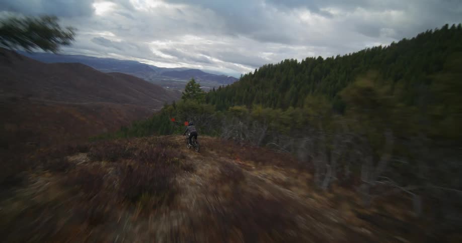 An aerial drone follows a mountain biker speeding down a narrow, winding trail on a rugged mountainside on a cloudy autumn day