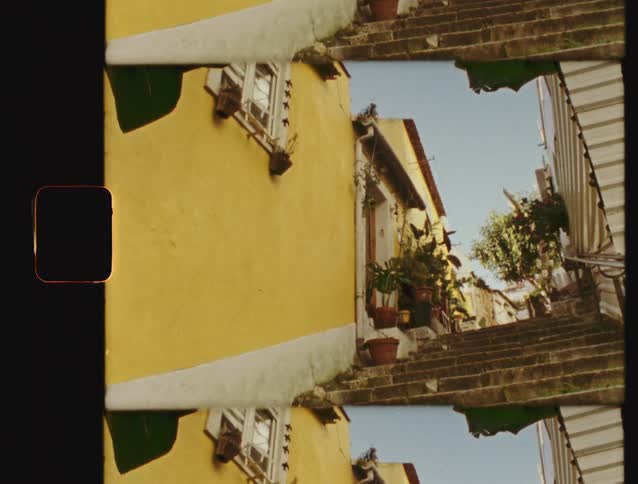 Vintage film emulation of pigeons flying up a narrow cobblestone staircase in a sunlit European alley with yellow buildings