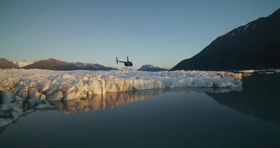 A helicopter takes off and flies low over a majestic Alaskan glacier and its reflective lake during a beautiful sunset