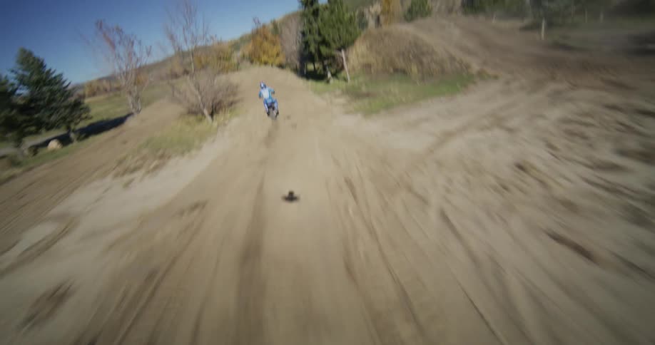 A fast FPV drone chases a motocross rider as they speed through jumps and turns on a dusty dirt track