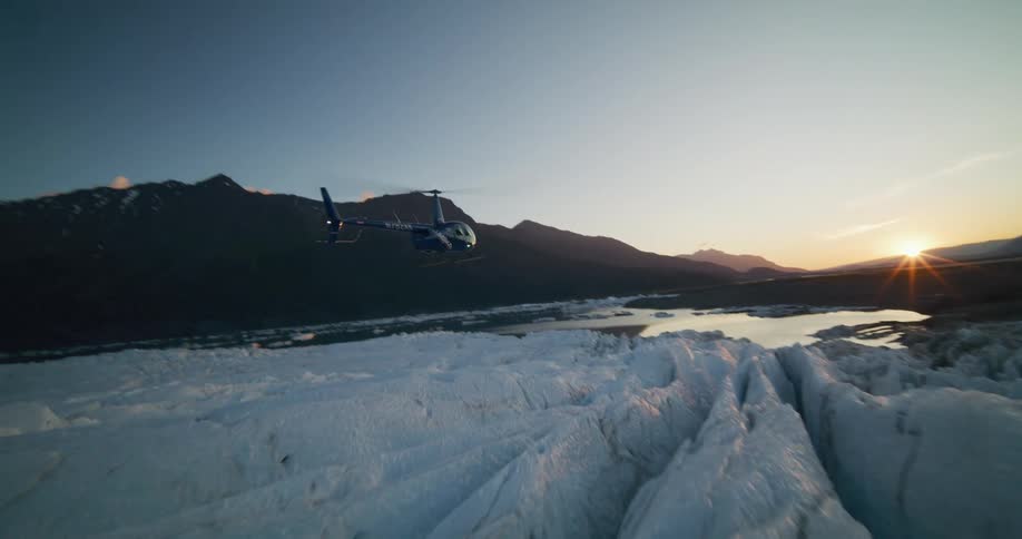 An epic aerial view of a helicopter taking off and flying over a vast, icy glacier in Alaska during a beautiful sunset