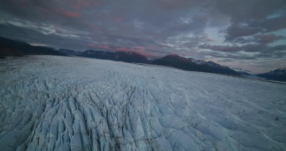 A breathtaking aerial shot slowly glides over a vast, cracked glacier field nestled between mountains during a dramatic sunset