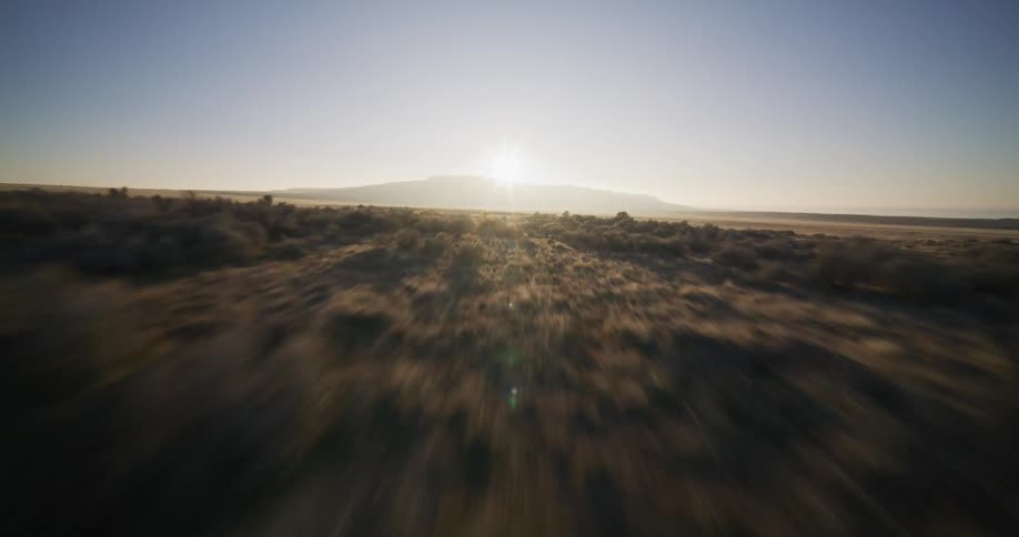 A dynamic, high-speed aerial flight soars over a vast, arid plain towards a distant mesa as the sun rises on the horizon