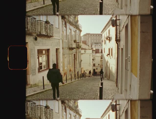 A Super 8 film scan shows a man walking down a charming, sunlit cobblestone street in an old European city